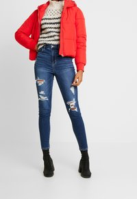 Person i röd puffjacka, krämfärgad och svart randig tröja, trasiga skinny jeans i blått och svarta klackstövlar som står på vit golv.