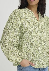 Blusa floreale verde con maniche a sbuffo, scollatura a V e polsini elastici. Tessuto di cotone con un dettagliato motivo botanico. Indossata con jeans.