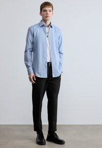 Helevalge pikkade varrukatega helesinine särk, mida kantakse avatud olekus valge t-särgi üle, kombineerituna mustade pükste ja mustade pits-up kingadega.