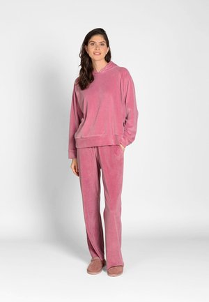 Woman standing wearing a matching soft pink velour hoodie and wide-legged pants with pink slippers against a plain white background.
