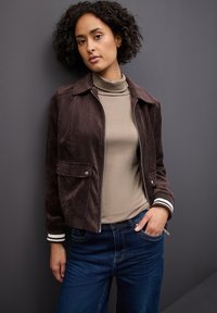Braune Cordjacke mit Reißverschluss, zwei Taschen und weißen gestreiften Bündchen, kombiniert mit einem beigen Rollkragenpullover und blauen Jeans.