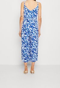 Donna che indossa un tuta senza maniche a fiori blu e bianchi con gambe larghe e sandali con tacco beige, vista di schiena contro un muro semplice.