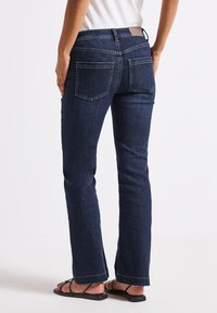 Jeans en denim bleu foncé avec une coupe droite, détails de couture visibles et une patch en cuir sur la taille. Portés avec des sandales noires.