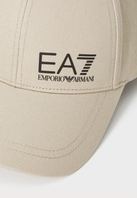 Căciulă de baseball bej, fabricată dintr-un material texturat, cu o visier�ă cusută și logo-ul negru EA7 Emporio Armani pe partea din față.