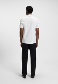Weißes kurzärmliges Poloshirt mit Kragen, kombiniert mit schwarzen Hosen, von hinten betrachtet. Einfarbiger Stoff ohne sichtbare Muster.