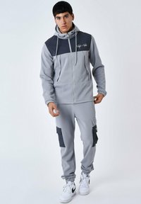 Felpa grigia con zip e pannello centrale grigio scuro, cappuccio regolabile e polsini elasticizzati, abbinata a joggers grigi coordinati con tasche cargo.
