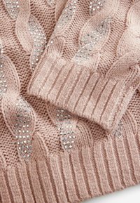 Kabelstrickpullover in sanftem Pink mit gerippten Bündchen, ausgestattet mit silbernen Verzierungen und einem strukturierten Muster.