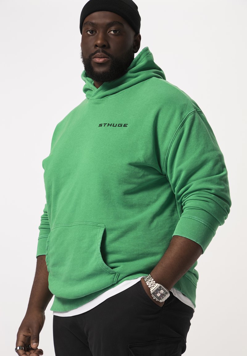 Felpa verde con tasca frontale e logo stampato in nero. Presenta un cappuccio con cordoncino e polsini a costine. Indossata sopra una maglietta bianca.