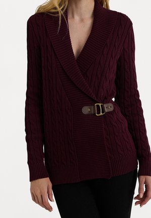 Femme portant un cardigan en maille torsadée bordeaux foncé avec un large col châle et une ceinture marron à boucle sur le côté, associé à un pantalon noir.
