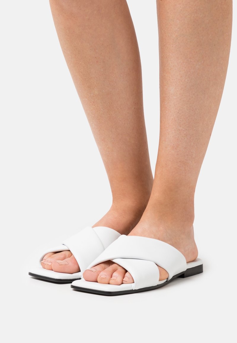Sandales plates blanches avec de larges straps croisées, texture lisse, forme carrée à l'avant, et semelle noire, portées pieds nus.