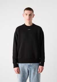 Sudadera negra con puños y dobladillo acanalados, que presenta un pequeño logotipo blanco de "Calvin Klein" en el pecho. Combinada con jeans de mezclilla azul claros.