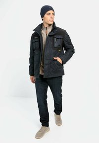 Sort polstret jakke med flere lommer og høj krave, båret over en beige sweater, parret med mørke jeans og lyse sneakers.