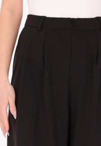 Pantaloni neri a vita alta con due pince frontali, tessuto morbido, passanti per cintura e una vita liscia. Vista ravvicinata della zona della vita.