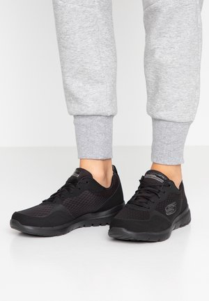 Czarne sneakersy Skechers noszone z jasnoszarymi spodniami dresowymi, pokazane na białej podłodze na jednolitym tle.