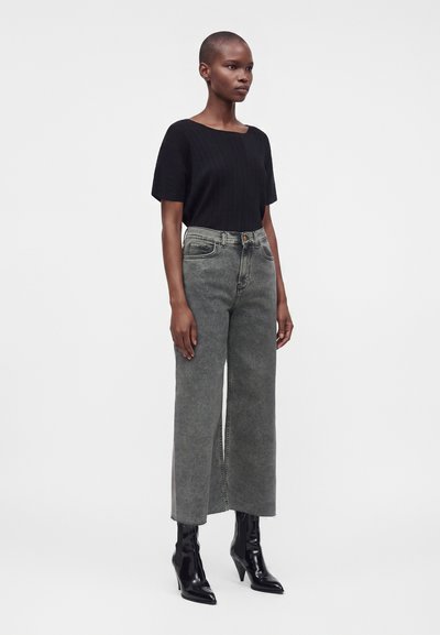 Femme aux cheveux courts portant un haut noir à manches courtes, un jean gris taille haute à jambes larges, et des bottes noires à talons, debout sur un fond blanc.