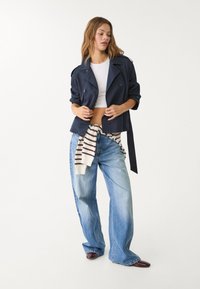 Marindubbelknäppt jacka, vit kort topp, överdimensionerade ljusa jeans och en randig tröja knuten runt midjan, kompletterad med bruna skor.