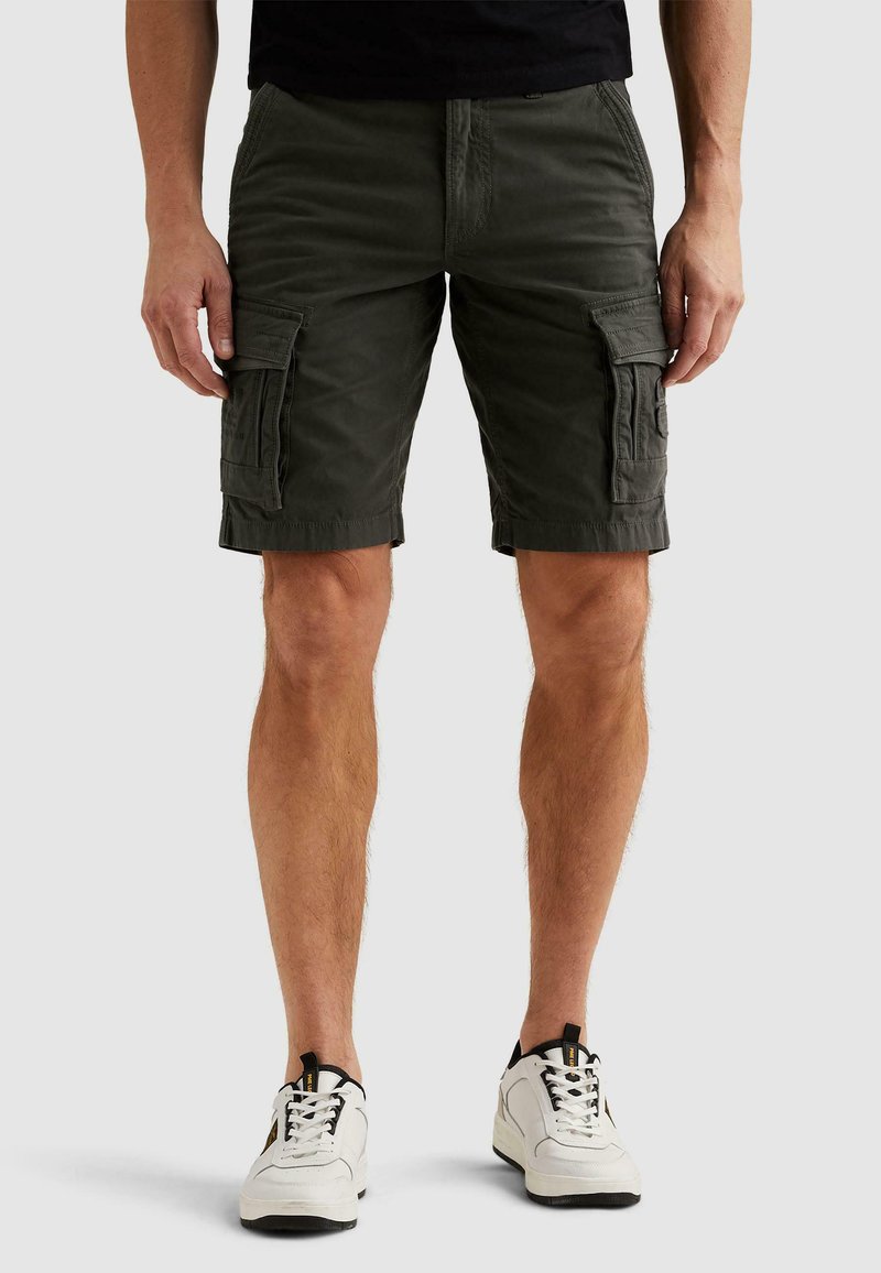 Homme portant un short cargo noir avec des poches latérales et des baskets blanches, debout devant un fond gris clair uni.