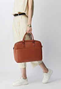 Maletín de piel marrón con dos asas, textura suave y un bolsillo con cremallera en el frontal. Combinado con pantalones crema y zapatillas blancas.