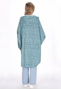 Hosszú, bő szabású kabát türkizkék színben, világos leopárdmintával, kapucnival és combközépig érő hosszúsággal, világoskék bőszárú farmerrel és fehér cipővel párosítva.