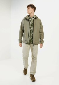 Leichte, olivgrüne Jacke mit Kapuze über einem gemusterten grünen Hemd, kombiniert mit beigen Hosen und braunen Sneakers, die vor einem weißen Hintergrund geht.