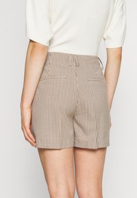Femme portant un short taille haute à carreaux marron et blanc et un haut crème à manches courtes, vue de dos.