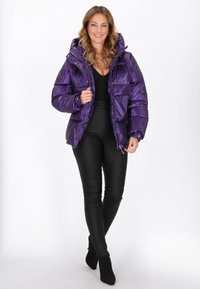 Lila puffertöltettel ellátott dzseki kapucnival, fényes textúrával és arany színű kiegészítőkkel. Fekete, testhezálló nadrág és magassarkú bokacsizma fölé van viselve.