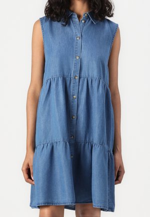 Ærmeløs blå denim kjole med knapper, lagdelt nederdel og spredt krave, båret af en stående person med krøllet hår.