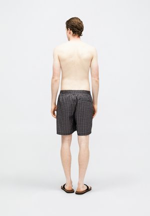 Bărbat stând desculț, fără cămașă, purtând șorturi în carouri alb-negru și papuci negri, pe un fundal simplu, deschis la culoare.