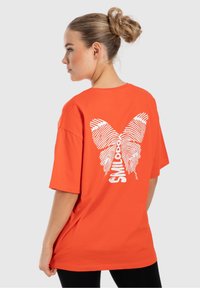Oranžové bavlněné tričko s oversized střihem, na zadní straně s designem bílého motýla a textu, krátké rukávy a kulatý výstřih.