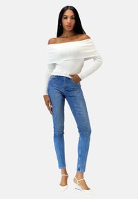 Hvid off-shoulder ribbet sweater, blå skinny jeans og hvide spidse hæle. Outfitet har en tætsiddende silhuet og tekstureret stof.