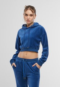 Felpa in velluto blu tagliata con cappuccio e cordini, caratterizzata da un discreto logo e polsini a coste. Abbinata a pantaloni jogger in velluto coordinati.