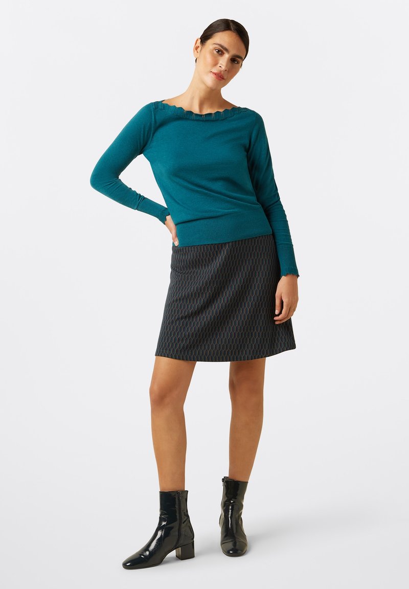 Teal sweater with scalloped neckline, paired with a dark patterned skirt. Model wearing black patent ankle boots, standing against a white background.