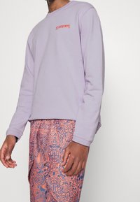 Persona che indossa una camicia a maniche lunghe lavanda con il testo "LES BENJAMINS" e pantaloni arancioni con un motivo elaborato.