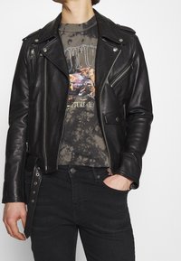 Černá kožená motorkářská bunda se stříbrnými zipy, knoflíkovými detaily a ramenními vycpávkami, nošená přes tričko s grafickým potiskem ve stylu tie-dye a černé džíny.