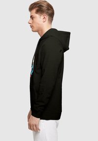 Svart zip-up hettegenser med frontlomme, med et fargerikt grafisk design på brystet. Mykt stoff, pullover-stil, og festet hette.
