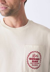 Camiseta de manga corta beige con un bolsillo en el pecho que presenta un logo circular rojo y el texto "Billy's Burger Bar" junto con los detalles de la dirección en blanco.