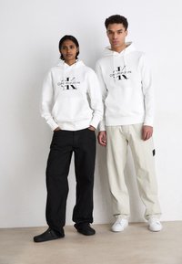 Bijele dukserice s crnim logom, crne i krem hlače te crne klompe. Manekenke stoje nasuprot neutralnoj pozadini.