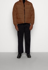Brązowa ocieplana kurtka puchowa o prostokątnym kroju, z wysokim kołnierzem i zapięciem na zamek błyskawiczny. Połączona z ciemnymi spodniami i czarnymi sneakersami.