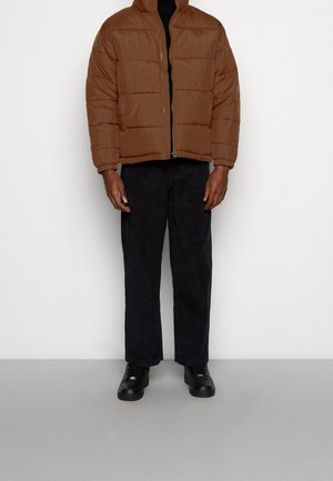 Braune isolierte Daunenjacke mit kastigem Design, hohem Kragen und Reißverschluss. Kombiniert mit dunklen Hosen und schwarzen Sneakers.