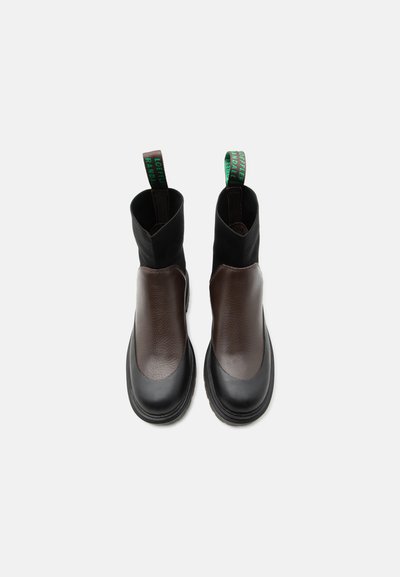 Ζευγάρι μαύρα και σκούρα καφέ δερμάτινα μποτάκια αστραγάλου με παχιά σόλα και πράσινες ταινίες για τράβηγμα με κείμενο σε λευκό φόντο.