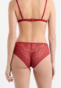 Conjunto de lencería de encaje rojo con un patrón en zigzag y bordes festoneados, con tirantes ajustables y textura transparente.