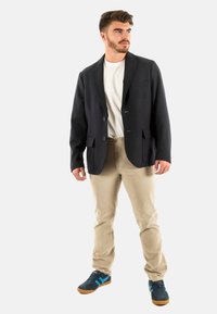 Blazer navy con rever a lancia, due tasche frontali e tre bottoni, abbinato a una T-shirt bianca, pantaloni beige e sneakers con bordini blu.
