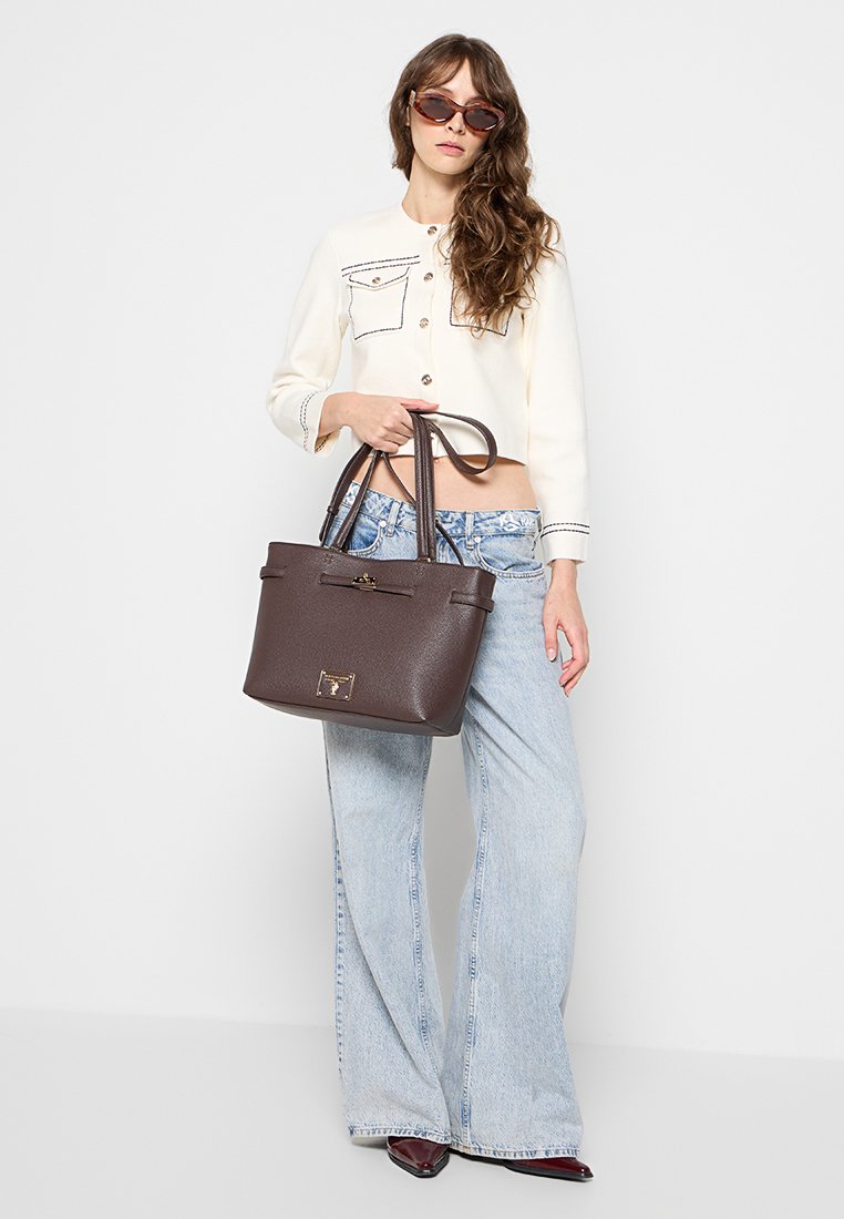 Bolso de mano de cuero marrón con doble asas, forma estructurada y detalle de cierre en la parte frontal. El modelo lleva puestos unos jeans acampanados de color azul claro y una blusa crema.