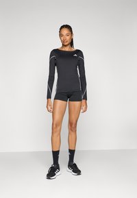 Črna atletska majica z dolgimi rokavi z belimi poudarki, kombinirana s črnimi kratkimi hlačami in črnimi atletskimi nogavicami. Model nosi črne tekaške copate.