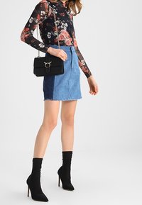 Langärmliges Blumen-Mesh-Top, blaue Jeans-Minirock mit rohem Saum, schwarze Handtasche mit Kettenriemen und schwarze knielange Stilettos.