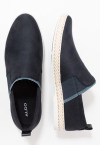 Scarpe slip-on blu navy con tomaia in suede morbido, suola bianca in gomma e dettagli intrecciati beige sul tallone e attorno alla suola.