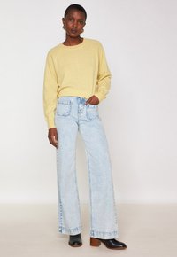 Donna che indossa un maglione giallo a maniche lunghe, jeans a gamba larga blu chiaro, stivali neri con tacco e orecchini a cerchio dorati, in piedi con una mano in tasca.