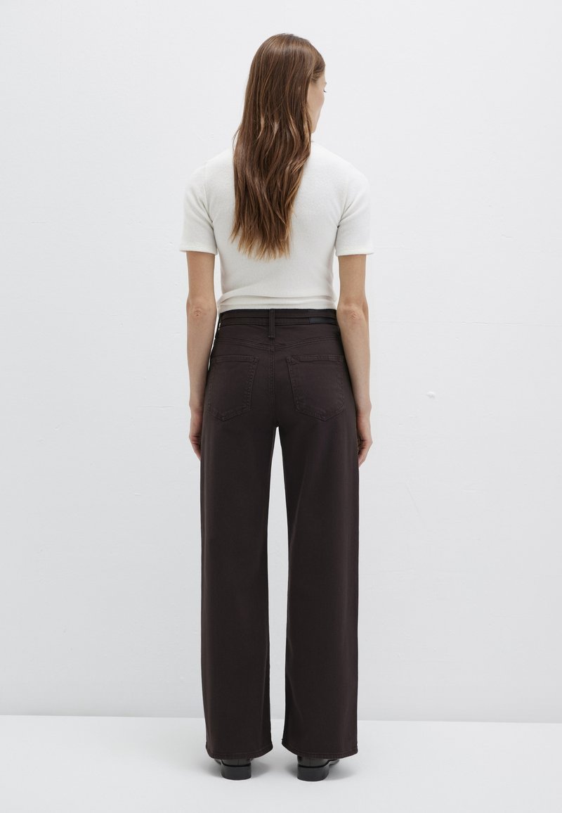 Femme aux longs cheveux bruns se tient face à un mur blanc, portant un haut blanc à manches courtes et un pantalon noir taille haute à jambes larges.