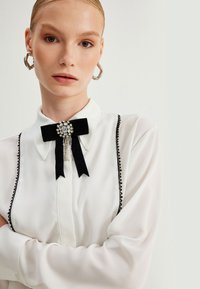 Blusa blanca con ribete negro y un lazo de terciopelo decorado con un broche de joyas. Cuenta con mangas largas y una textura suave.