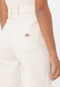 Pantalones blancos de talle alto hechos de mezclilla con un acabado texturizado. Presentan un bolsillo de parche con un colorido logo de Dickies en la parte trasera.